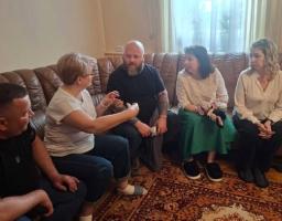 Група з п'яти людей сидить на великому коричневому шкіряному дивані та поруч із ним у кімнаті з килимом на підлозі, активно спілкуючись між собою, при цьому жінка у білій футболці на передньому плані жестикулює руками