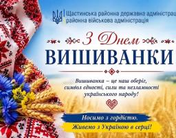 Святкова листівка від Щастинської РДА на тлі блакитного неба та пшеничного поля містить великий привітальний напис «З Днем ВИШИВАНКИ!», патріотичні гасла про вишиванку як оберіг та букет синіх волошок і колосків, що лежить на біло-червоному вишитому рушнику.