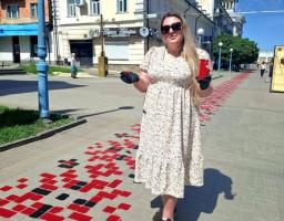 Молода жінка у світлій сукні в дрібний квіточку, сонцезахисних окулярах та чорних рукавичках стоїть посеред пішохідної вулиці міста поруч із намальованим на бруківці великим червоно-чорним орнаментом вишиванки, тримаючи в руках паперовий стаканчик.
