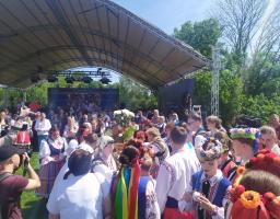 На зображенні представлено масовий захід просто неба біля великої критої сцени, де зібралося багато людей, частина з яких вбрана в традиційні українські вишиванки, вінки з барвистими стрічками та народні костюми