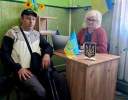 У кабінеті із зеленими стінами чоловік у кепці та з милицями сидить на стільці поруч із дерев'яним робочим столом, за яким сидітиме жінка в окулярах та червоному піджаку на тлі державного прапора України.