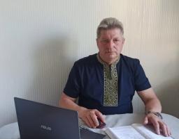 Чоловік середнього віку із сивим волоссям у темно-синій сорочці з вишитим коміром та планкою сидить за столом перед відкритим ноутбуком ASUS, тримаючи руку на комп'ютерній миші поруч із розгорнутим блокнотом