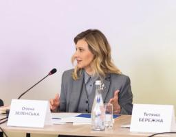 Фотографія Олени Зеленської у сірому костюмі-трійці, яка сидить за робочим столом із мікрофоном, документами та табличками з іменами й активно виступає, жестикулюючи руками під час офіційного заходу