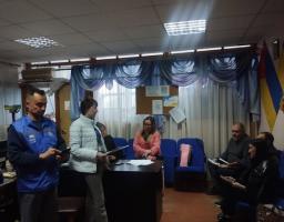 У кабінеті з великим вікном, завішеним блакитним тюлем, та українським прапором на стіні зафіксовано робочу зустріч кількох людей, де частина присутніх сидить у синіх кріслах, а жінка у світлій куртці та чоловік у синьому жилеті УВКБ ООН стоять біля письмового столу