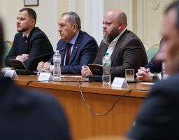 Три посадовці в ділових костюмах сидять за довгим дерев'яним столом президії з мікрофонами та пляшками води під час офіційного засідання, а на передньому плані видно розмиті силуети інших учасників заходу