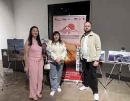Дві жінки та чоловік стоять у виставковій залі на тлі вертикального банера з написом «Актуальні голоси Луганщини», оточені розміщеними на мольбертах-триногах світлинами з краєвидами та зруйнованими будівлями.