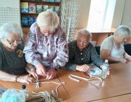 Чотири літні жінки сидять та стоять за великим дерев'яним столом у світлій кімнаті, займаючись рукоділлям із використанням ниток, стрічок та ножиць