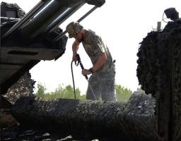 Військовослужбовець у камуфляжній формі та панамі миє артилерійську установку водою зі шланга високого тиску просто неба на тлі дерев