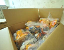 Велика картонна коробка, наповнена продуктовими наборами гуманітарної допомоги в прозорих пластикових пакетах з автентичними продуктами, ложками та консервами, що стоїть у приміщенні на тлі інших складених коробок