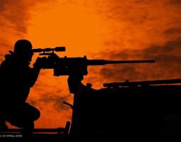 Силует військовослужбовця, який цілиться крізь оптичний приціл великокаліберного кулемета на тлі яскравого помаранчевого неба під час заходу або сходу сонця