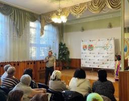 Чоловік із мікрофоном виступає на сцені залу перед сидячою авдиторією слухачів, а праворуч від нього встановлено інформаційний стенд із написом «Луганщина моя».