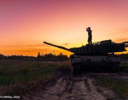 Силует військового танка з бійцем на башті, який дивиться вглиб поля на тлі яскравого помаранчево-рожевого заходу сонця.