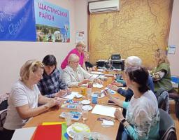 Група переважно літніх жінок сидить за довгим столом у кабінеті під час творчого заняття з малювання, де на стінах розміщені плакат «Щастинський район» та велика старовинна мапа