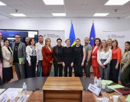 Група людей стоїть у конференційній залі для спільного фото на тлі прапорів України та Європейського Союзу, інформаційних стендів Міністерства соціальної політики та великого екрана