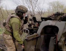 Український військовослужбовець у камуфляжній формі та шоломі з навушниками говорить по рації, перебуваючи на бойовій позиції біля великої артилерійської установки серед дерев