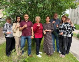 Група з восьми людей стоїть у шеренгу на фоні зеленого дерева біля будівлі та тримає в руках патріотичні синьо-жовті сувеніри або дзвіночки на стрічках.