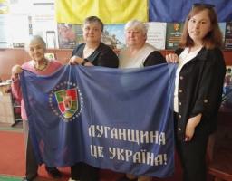 Колаж із чотирьох фотографій демонструє патріотичний захід просто неба, де група людей похилого та молодого віку позирує біля виставкових стендів із фотографіями, прапором Луганщини, виробами ручної роботи та банером із написом «Луганщина — це Україна»