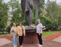 Дві жінки та чоловік стоять біля підніжжя високого бронзового пам'ятника ангелу з німбом і довгим списом, де чоловік кладе м'яку іграшку ведмедика поруч із квітами на знак пам'яті