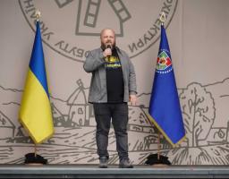 Чоловік із мікрофоном у руках виступає на сцені між державним прапором України та прапором Луганської області на тлі банера з традиційними українськими мотивами
