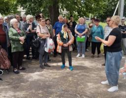 Група людей різного віку зібралася півколом на подвір'ї в тіні дерев, уважно слухаючи жінку в чорній футболці та широких джинсах, яка стоїть праворуч і жестикулює під час розмови.