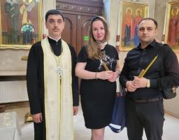 Православний священник у чорному підряснику та світлій єпітрахілі стоїть у храмі поруч із жінкою та чоловіком у темному одязі, які тримають у руках тонкі церковні свічки на тлі ікон та вхідних дверей.