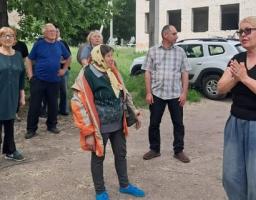 Група людей різного віку стоїть на вулиці біля цегляної будівлі та автомобіля, слухаючи жінку в окулярах та чорній футболці, яка щось емоційно розповідає та тримає руки разом