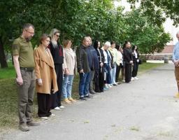 Група людей вишикувалася в шеренгу на асфальтованій доріжці в парку, схиливши голови під час хвилини мовчання, тоді як навпроти них стоїть чоловік у блакитній сорочці та штанах карго