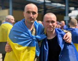 Два звільнених з полону українських військовослужбовці з поголеними головами дружньо обіймаються за плечі на вулиці, загорнувшись у синьо-жовті прапори України