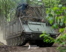 Гусеничний бронетранспортер М113, додатково захищений металевими решітками та маскувальною сіткою «мангал» на даху, рухається ґрунтовою дорогою серед лісового масиву, а з люка визирає водій-механік