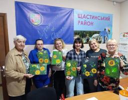 У приміщенні шість жінок різного віку стоять у ряд перед столом і тримають у руках свої малюнки з яскравими соняшниками на тлі прапора з написом «Луганщина це Європа» та плаката «Щастинський район»