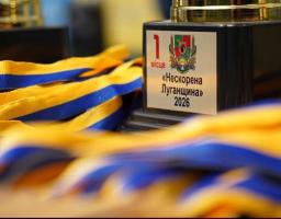 Спортивний кубок за перше місце з гербом Луганської області та написом «1 місце «Нескорена Луганщина» 2026» стоїть на столі поруч із купою розмитих на передньому плані синьо-жовтих нагородних стрічок.