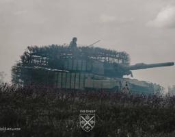 На фото зображено український танк Leopard 2 з масивним захисним навісом («мангалом») від дронів та блоками динамічного захисту, що стоїть у полі під хмарним небом
