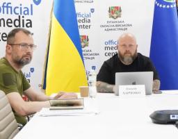 Двоє чоловіків сидять за білим столом під час засідання в офіційному медіацентрі Луганської обласної військової адміністрації на тлі прапорів України та відомства