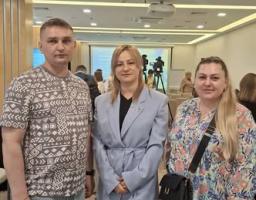 На фотографії троє людей — чоловік у футболка з візерунком та дві жінки у діловому й повсякденному вбранні — стоять поруч і посміхаються в конференц-залі.