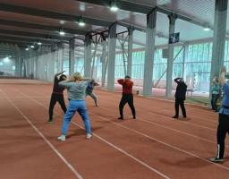 У великому критому легкоатлетичному манежі з біговими доріжками група людей старшого віку виконує фізичні вправи з розминки під керівництвом інструктора