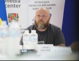На тлі банера Луганської обласної військової адміністрації за столом із пляшками води сидить лисий чоловік із густою бородою у темній футболці, уважно дивлячись убік