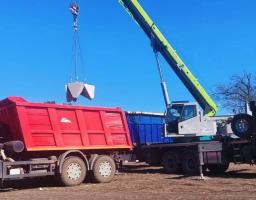 На тлі ясного синього неба автомобільний кран вантажить бетонні оборонні блоки («зуби дракона») у кузов великої червоної вантажівки, на даху кабіни якої стоїть чоловік