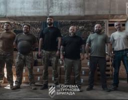 На світлині у закритому приміщенні стоять у ряд шестеро чоловіків у військовому та повсякденному одязі на тлі картонних коробок і дров, а внизу та у верхньому правому куті розміщено емблему з написом «ІІІ ОКРЕМА ШТУРМОВА БРИГАДА».