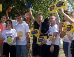 На тлі зелених дерев група молодих волонтерів у білих футболках радісно позирує для фото, тримаючи в руках жовті листівки з написом «Всесвітній день прибирання», круглі знаки-смайли та велику портативну аудіоколонку
