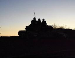 На зображенні на тлі вечірнього неба у світлі західу сонця видно темні силуети військового танка та кількох бійців, які сидять на його броні.