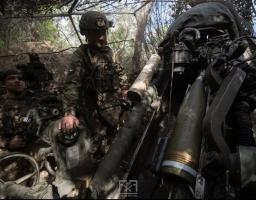 На зображенні під маскувальною сіткою в лісі двоє українських військових готують до пострілу артилерійську установку, великий снаряд якої завантажено в механізм.