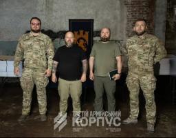 На фото зображено чотирьох чоловіків, двоє з яких у військовій формі камуфляжу «мультикам» та двоє у цивільному одязі мілітарі-стилю, які стоять у ряд у приміщенні на тлі прапора із зображенням лева та фортеці.