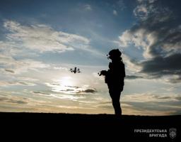 На зображенні на тлі вечірнього підсвіченого сонцем неба видно темний силует військового, який керує квадрокоптером, що летить поруч, а у нижньому правому куті розміщено логотип та напис «ПРЕЗИДЕНТСЬКА БРИГАДА»
