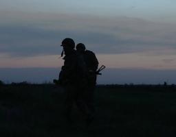 На фото зображено темні силуети двох військових у касках та зі спорядженням, які йдуть полем на тлі вечірнього або ранкового неба у сутінках.