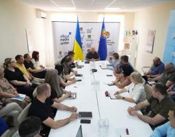 На фото зображено офіційне засідання або пресконференцію, де група людей сидить навколо довгого білого столу в кабінеті з українською символікою та прапорами