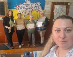 Група з п’яти жінок різного віку позує в кімнаті на тлі прапора та мапи України, при цьому троє з них тримають у руках грамоти, одна — пучок колосків, а жінка на передньому плані робить це селфі-фото.