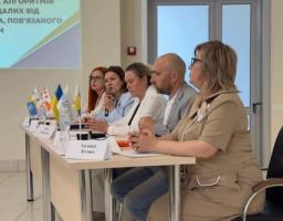 П'ятеро спікерів сидять за довгим столом із настільними прапорцями під час конференції, присвяченої допомозі постраждалим від насильства, пов'язаного з конфліктом.