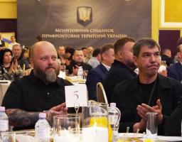 У великій залі на фоні банера з написом «Молитовний сніданок прифронтових територій України» за круглими сервірованими столами сидять численні учасники заходу, серед яких на передньому плані виділяються двоє чоловіків у чорному одязі, які аплодують.