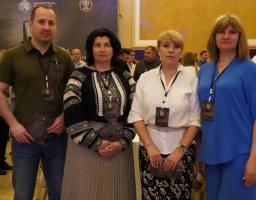 Один чоловік у зеленій футболці поло та три жінки в офіційно-діловому та традиційному одязі з бейджами на шиях стоять у ряд і позують для фото у великій залі на тлі інших людей та банера.