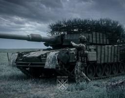 На фоні похмурого неба посеред зеленого поля стоїть замаскований гілками та блоками динамічного захисту військовий танк, біля якого спиною до глядача рухається солдат у повному бойовому спорядженні